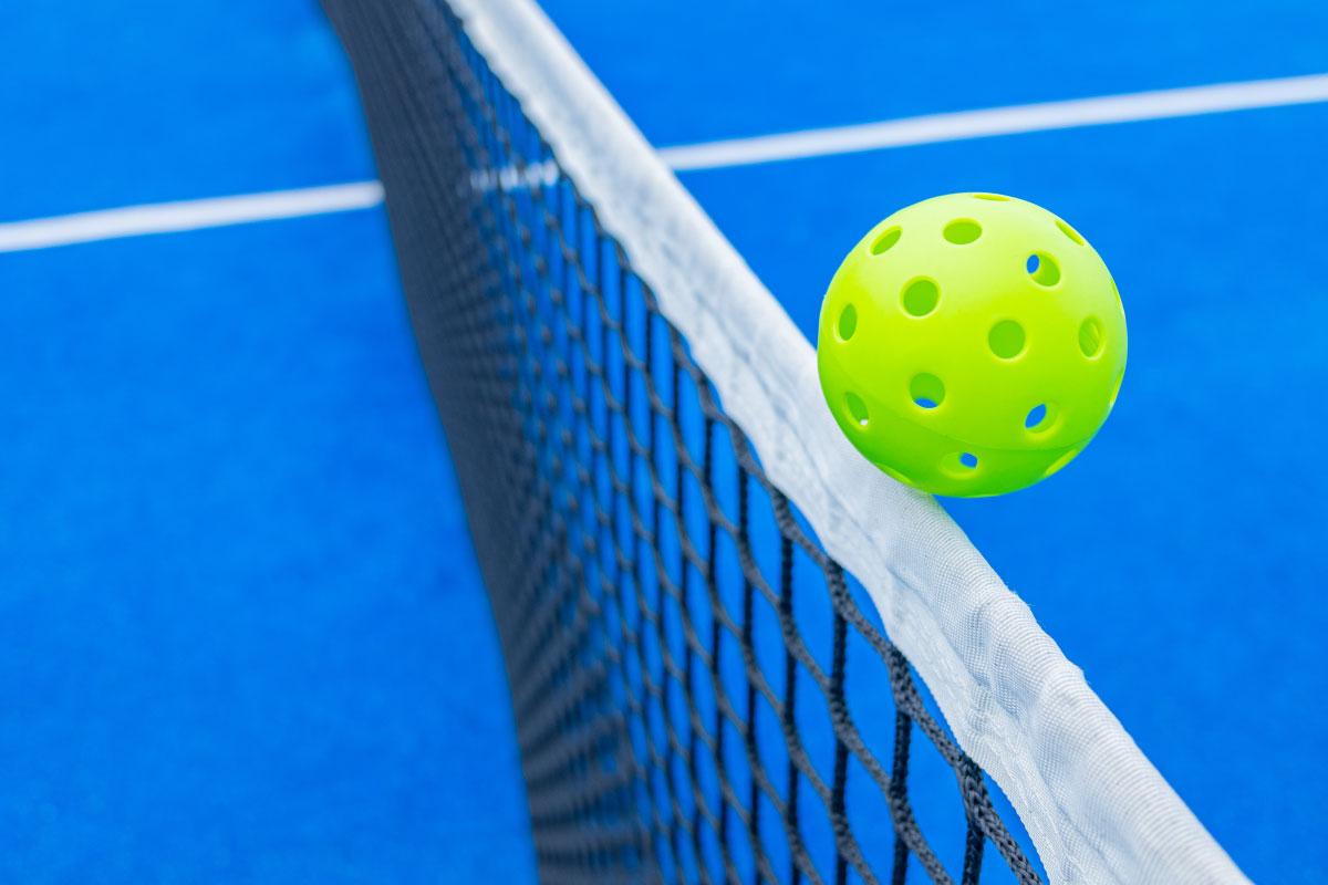 A bright yellow pickleball grazing over a net on a blue court.